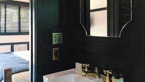 Une salle de bain avec un lavabo blanc, des robinets dorés et un miroir à motifs géométriques. Il y a deux interrupteurs sur le mur et une fenêtre avec des stores.