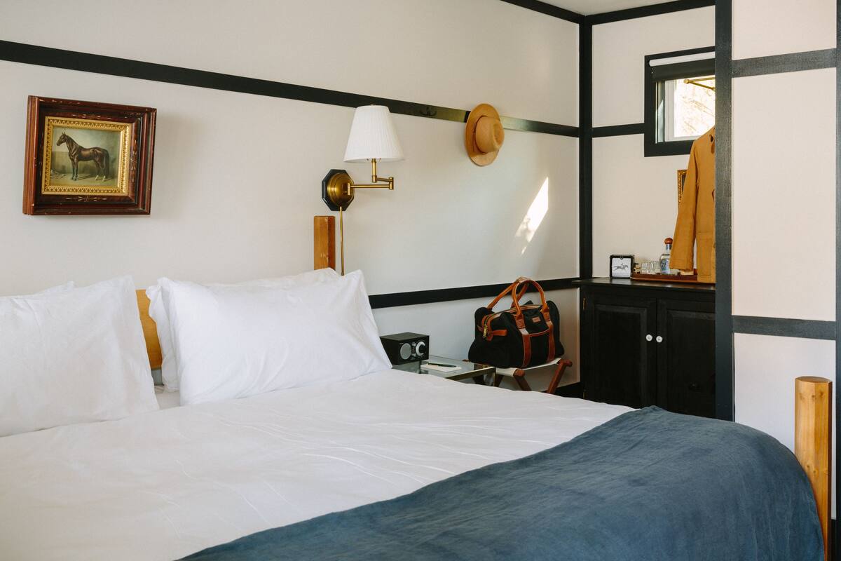 A hotel room with a bed, a nightstand, a lamp, a hat on the wall, a framed picture, and a wooden chair.