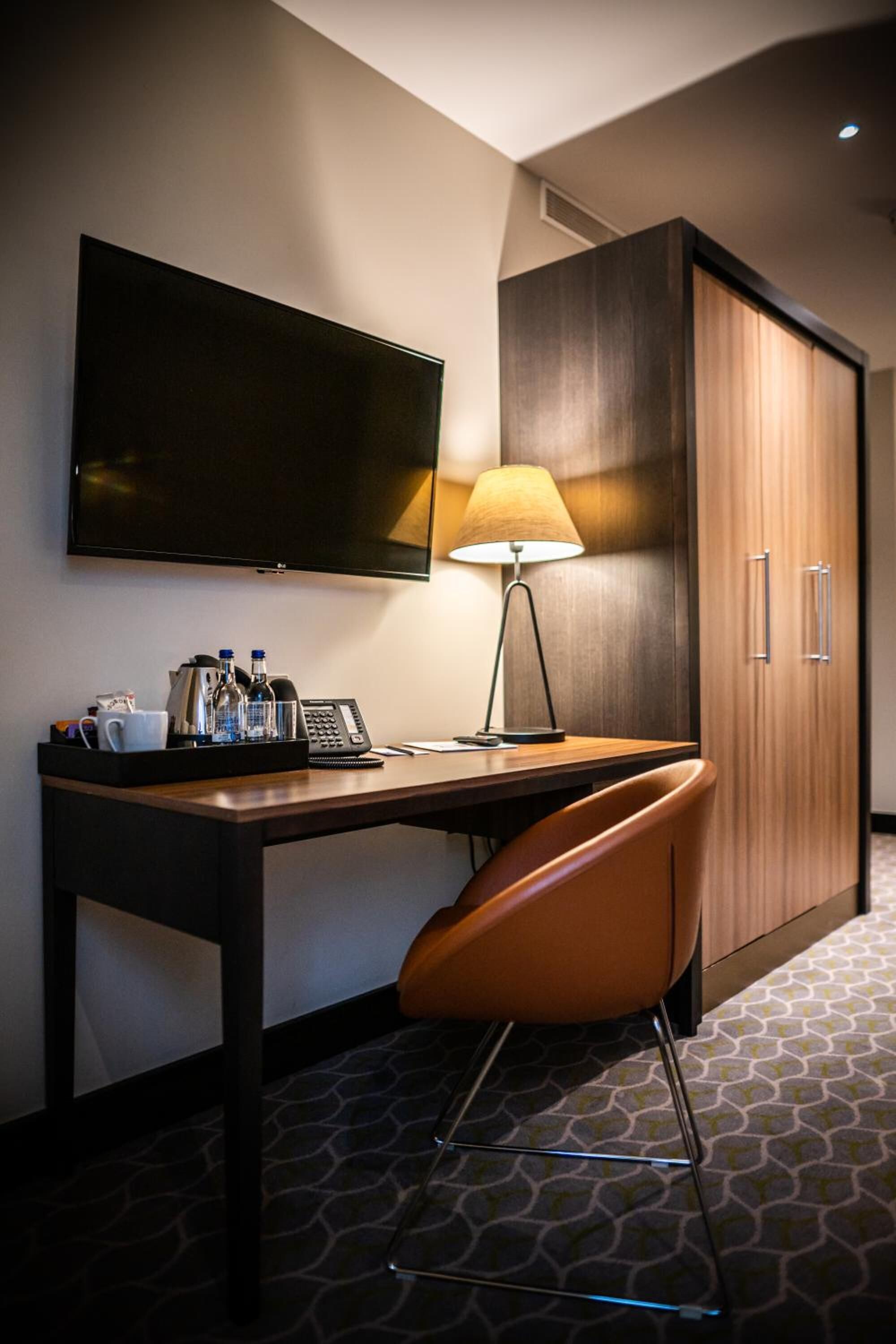 A hotel room with a desk, a chair, a television, a lamp, a minibar, and a wardrobe.