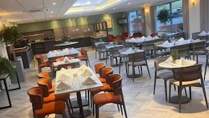 A spacious dining area with multiple tables and chairs, a kitchen in the background, and a modern ceiling design. - The Warwickshire Hotel and Country Club (Warwick)