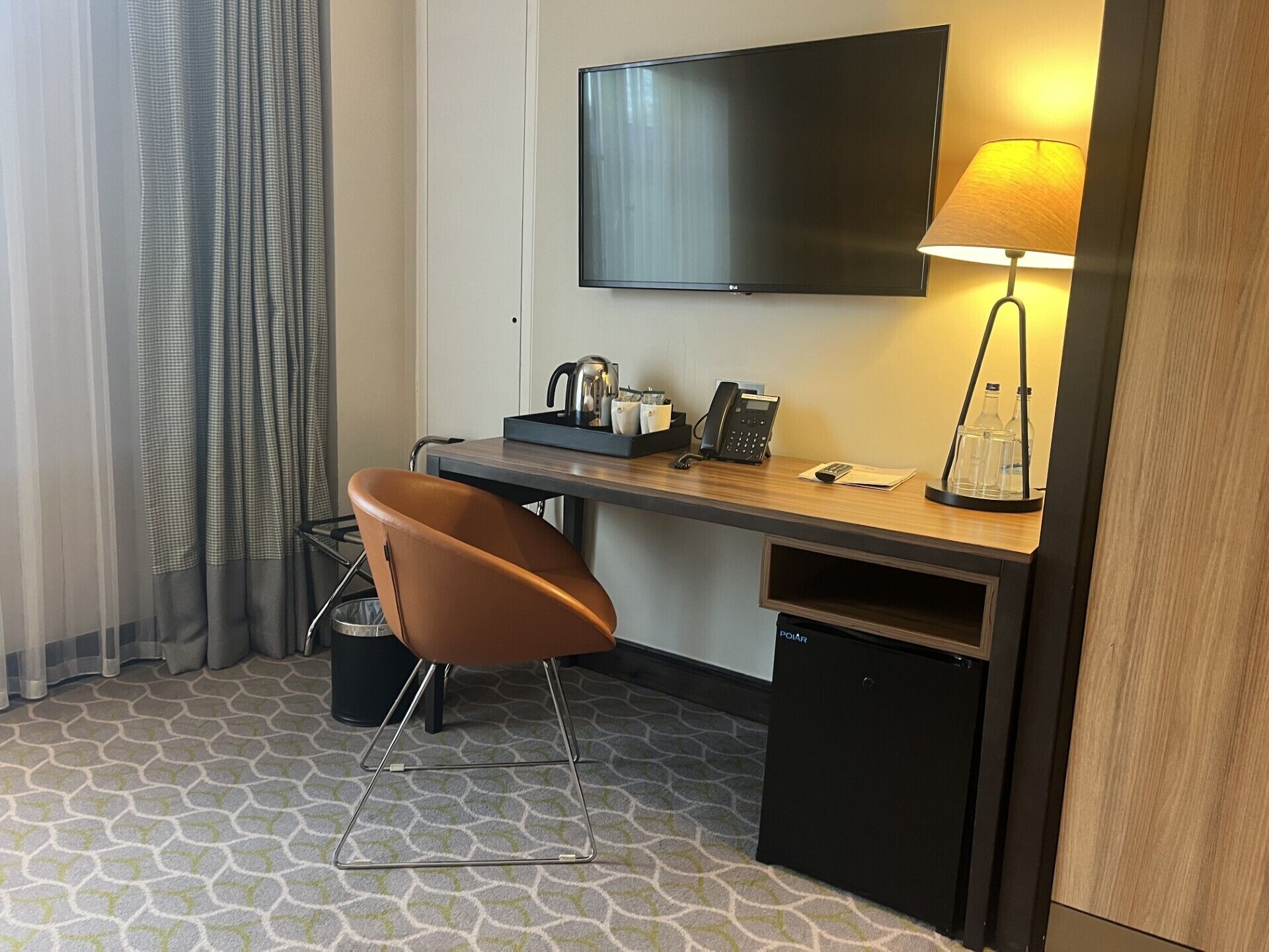 A hotel room with a desk, chair, television, lamp, and a trash bin.