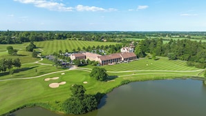 A golf course with a clubhouse, a pond, and surrounding greenery. - The Warwickshire Hotel and Country Club (Warwick)