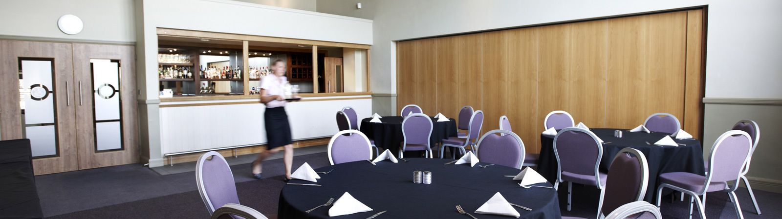 Un salón de conferencias con mesas redondas, sillas moradas y una zona de bar al fondo.