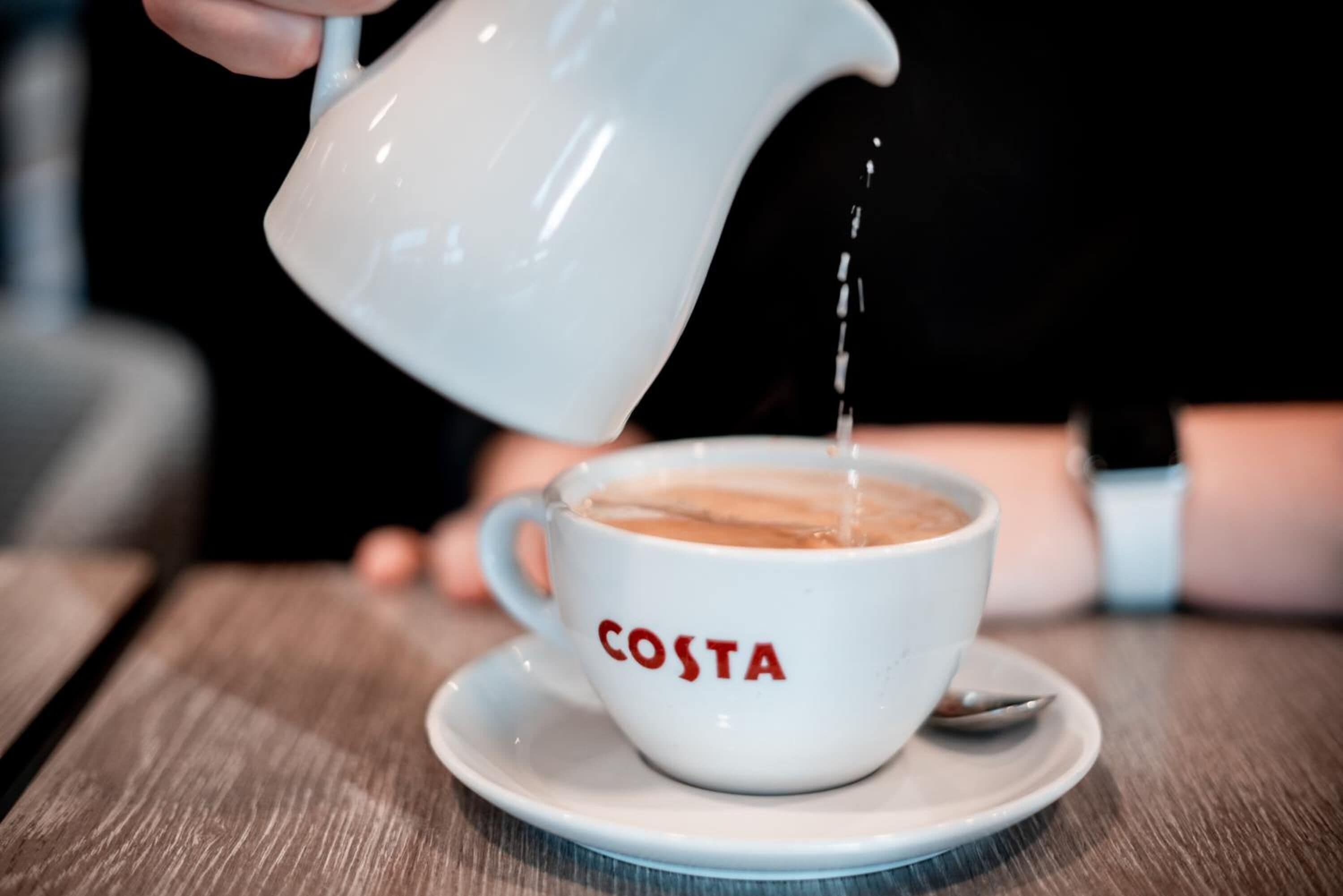 A white cup with "COSTA" written on it, being filled with a liquid from a white pitcher.