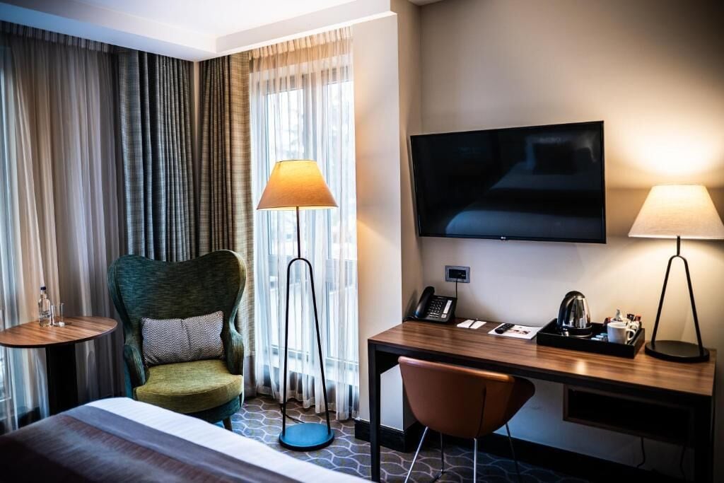 A hotel room with a bed, a desk with a chair, a television, a lamp, and a window with curtains.
