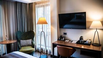 A hotel room with a bed, a desk with a chair, a television, a lamp, and a window with curtains.