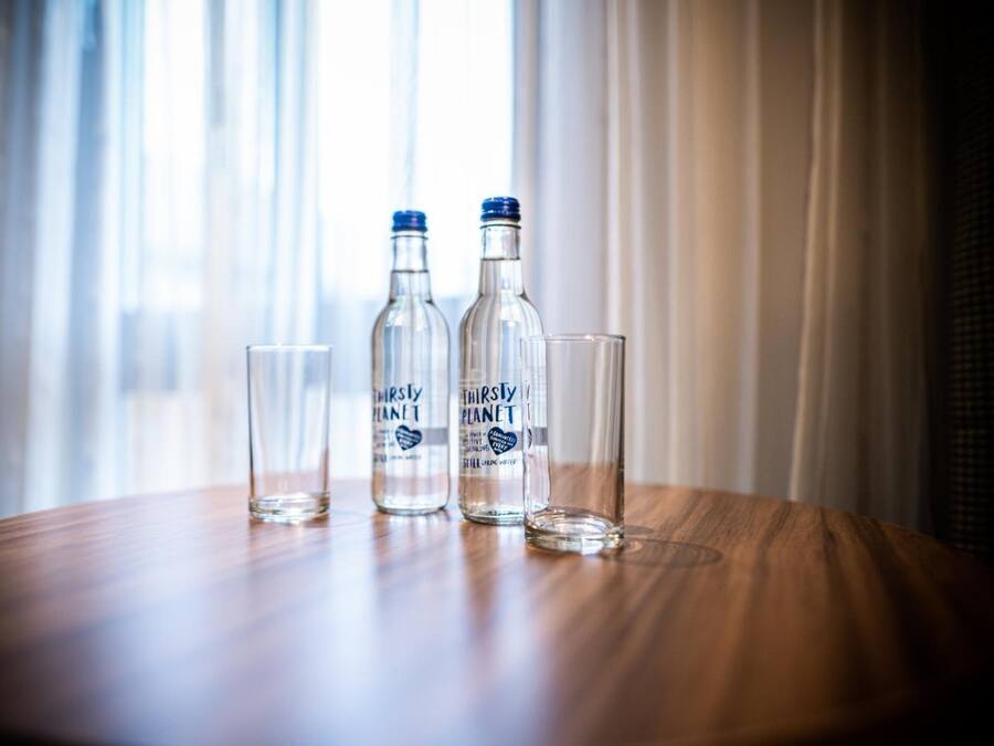 Drei klare Glasflaschen und zwei leere Gläser auf einem Holztisch neben einem Fenster mit durchsichtigen Vorhängen.