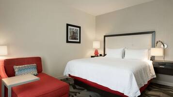A hotel room with a bed, a red armchair, a small table, and a nightstand with a lamp.