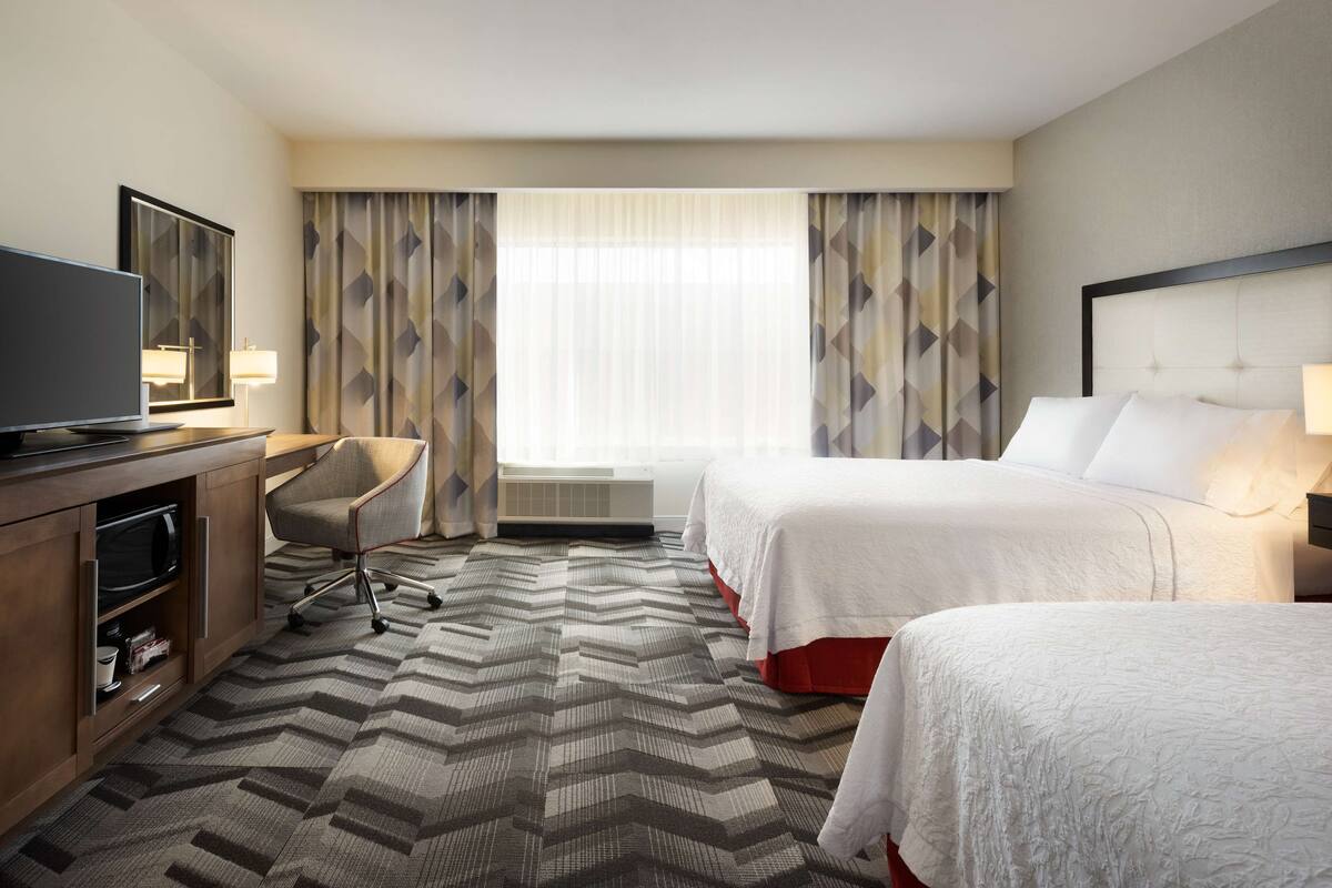 A hotel room with two beds, a desk with a TV, a chair, and a large window with curtains.