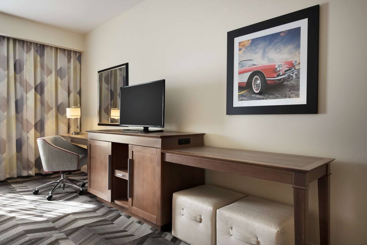A hotel room with a wooden desk, a television, a cushioned bench, a patterned curtain, and a framed picture of a car.