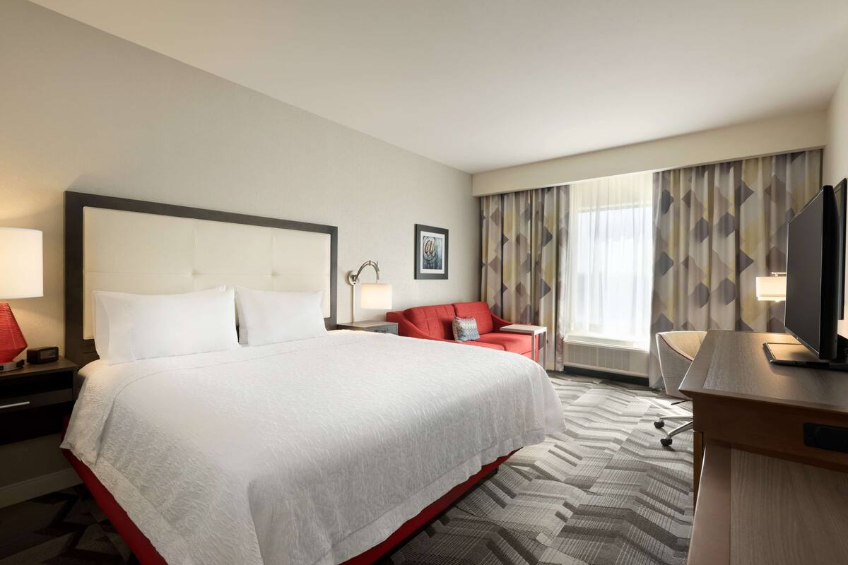 A hotel room with a large bed, a red armchair, a desk with a TV, and a window with curtains.