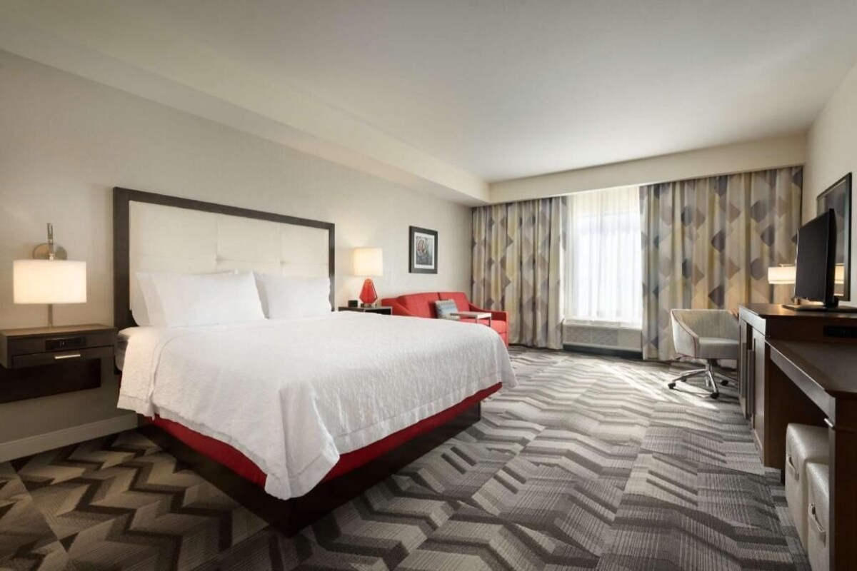 A hotel room with a large bed, a desk, a chair, a TV, and a window with curtains.