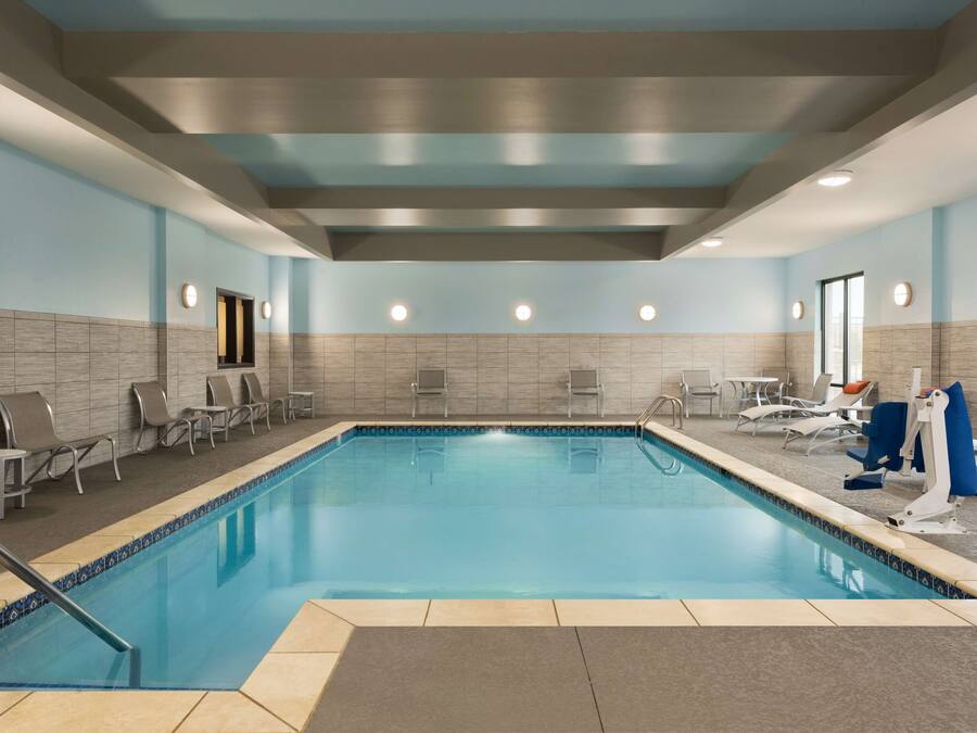 A swimming pool with lounge chairs and sun loungers on the sides, a ceiling with recessed lighting, and a tiled wall with a door.