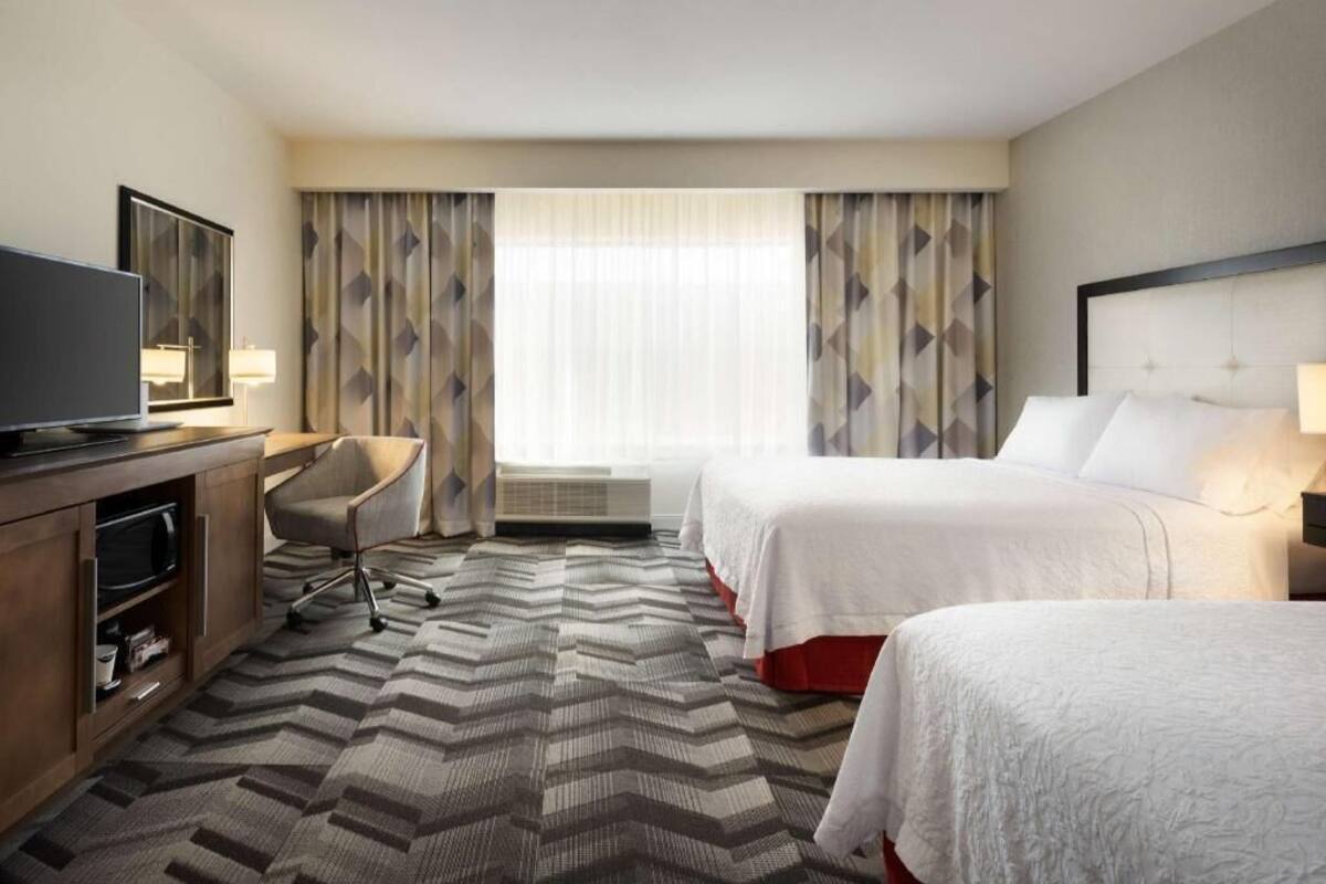 A hotel room with two beds, a desk, a TV, and a large window with curtains.