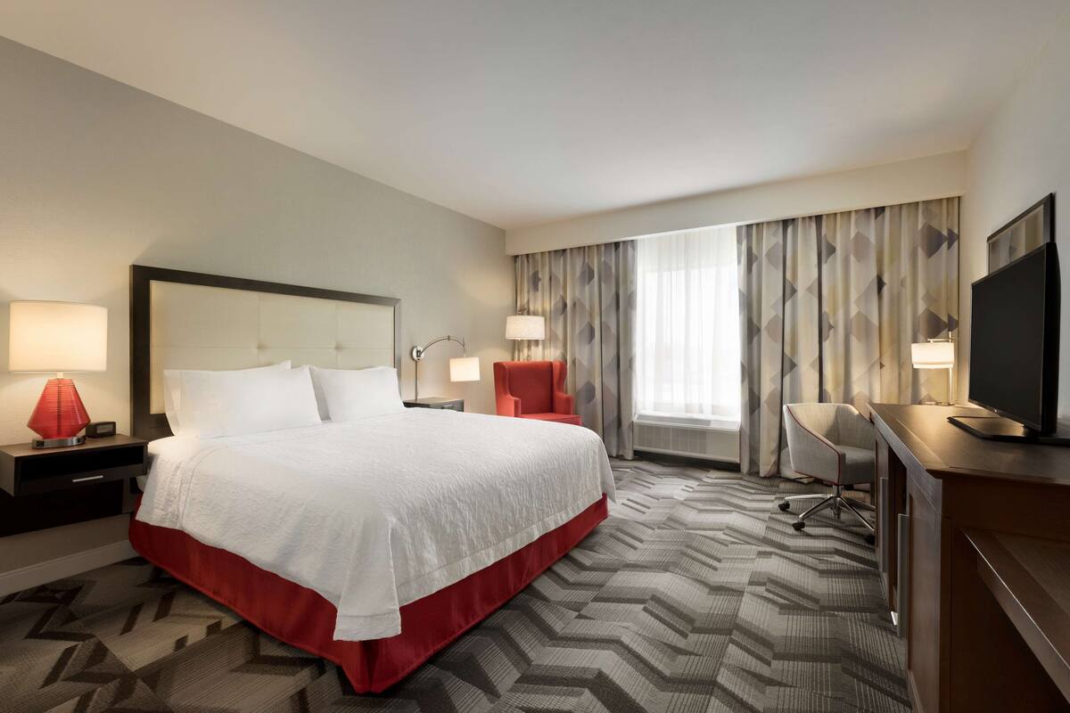 A hotel room with a large bed, a desk, a chair, a TV, and a window with curtains.