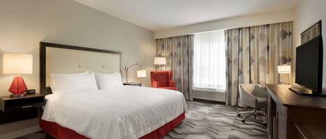 A hotel room with a large bed, a desk, a chair, a TV, and a window with curtains.