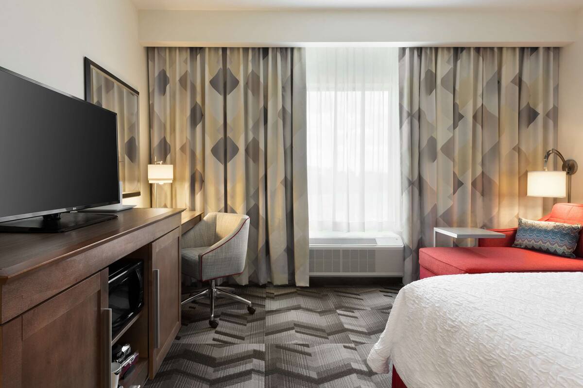 A hotel room with a flat-screen TV, a desk with a lamp, a red sofa, and a bed with a white comforter.