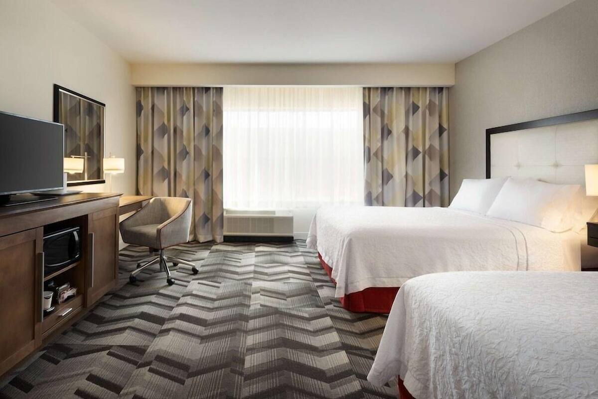 A hotel room with two beds, a desk with a television, a chair, and a large window with curtains.