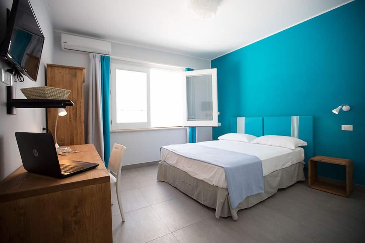 Un dormitorio con cama, un escritorio con laptop, un televisor montado en la pared y un ventanal con cortinas.