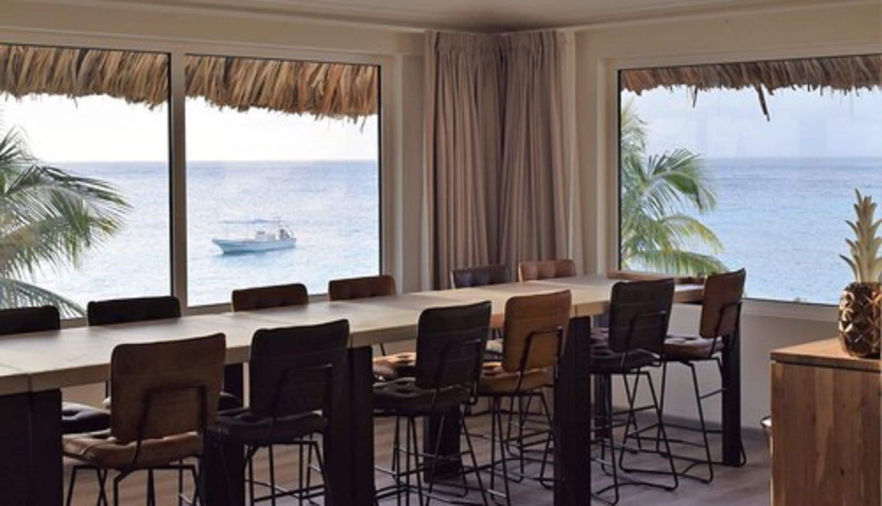 A dining area with a view of the sea, featuring a long table and several chairs.