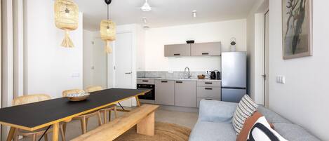 Una cocina moderna con encimera negra, armarios blancos y horno integrado. Hay una zona de comedor con mesa y sillas de madera, y un sofá gris con alfombra tejida.