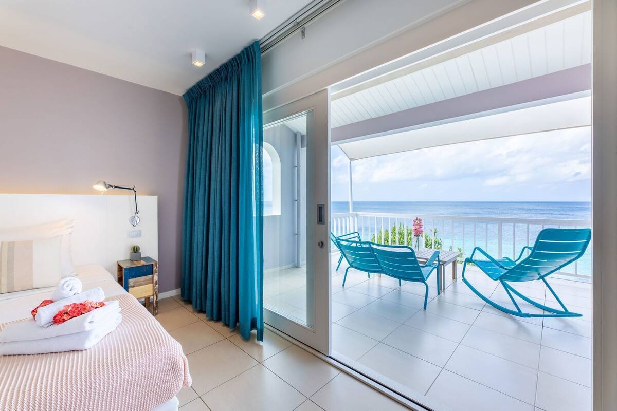 Une chambre d’hôtel comprenant un lit, une table de chevet, un bureau, une chaise, une table, un rideau et un balcon avec vue sur l’océan.