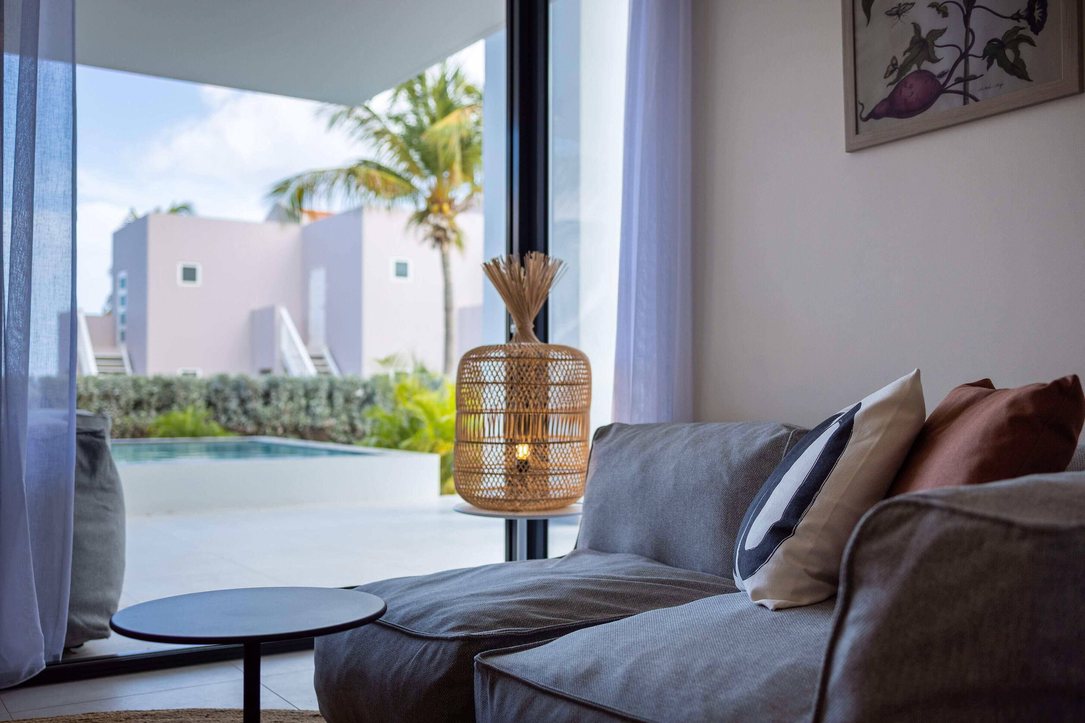 Un salón moderno con un sofá gris, una mesita redonda y una lámpara de mimbre. Un ventanal grande ofrece vistas a una piscina y palmeras.