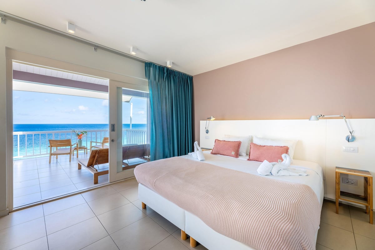 Une chambre d’hôtel avec un grand lit, une table de chevet, une vue sur l’océan et un balcon équipé de chaises et d’une table.