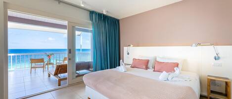 Habitación de hotel con una cama grande, una mesita de noche, vista al océano y un balcón con sillas y una mesa.