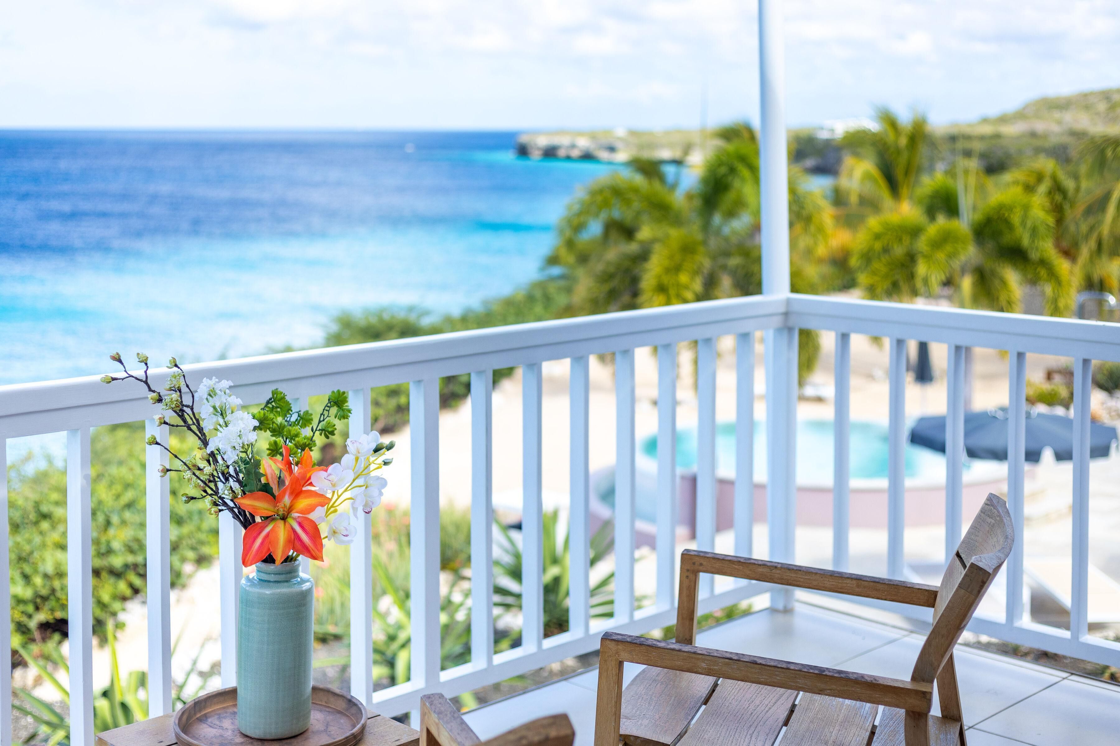 Une chaise en bois sur un balcon, avec vue sur l’océan, et un vase rempli de fleurs.