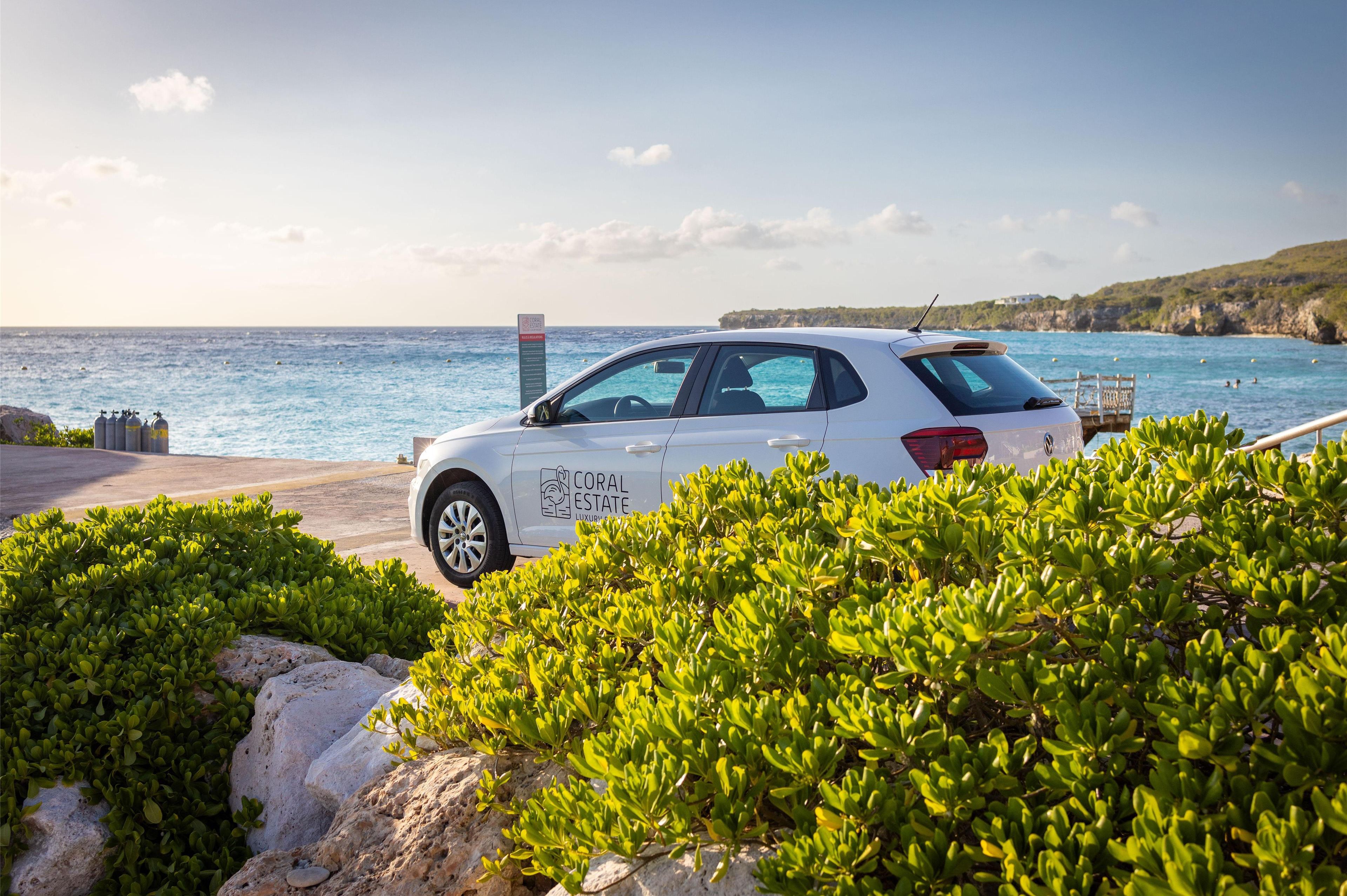A white car with "CORAL ESTATE" on the side parked near a coastal area with clear skies and greenery.