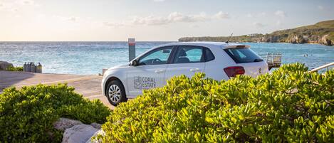 A white car with "CORAL ESTATE" on the side parked near a coastal area with clear skies and greenery.