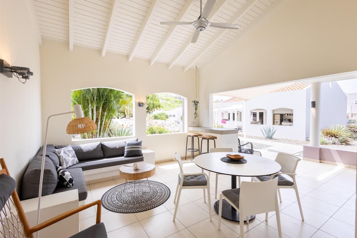 Un espace de vie lumineux et décloisonné, comprenant un canapé, une table à manger et des chaises, de grandes fenêtres offrant une vue sur la verdure et un ventilateur de plafond.