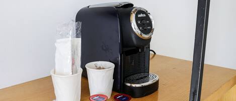 A coffee maker on a wooden table with a white cup, a plastic bag, and two small containers.