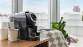 Une machine à café posée sur une table en bois, avec deux gobelets jetables, une fenêtre donnant sur des immeubles et une plante en arrière-plan.