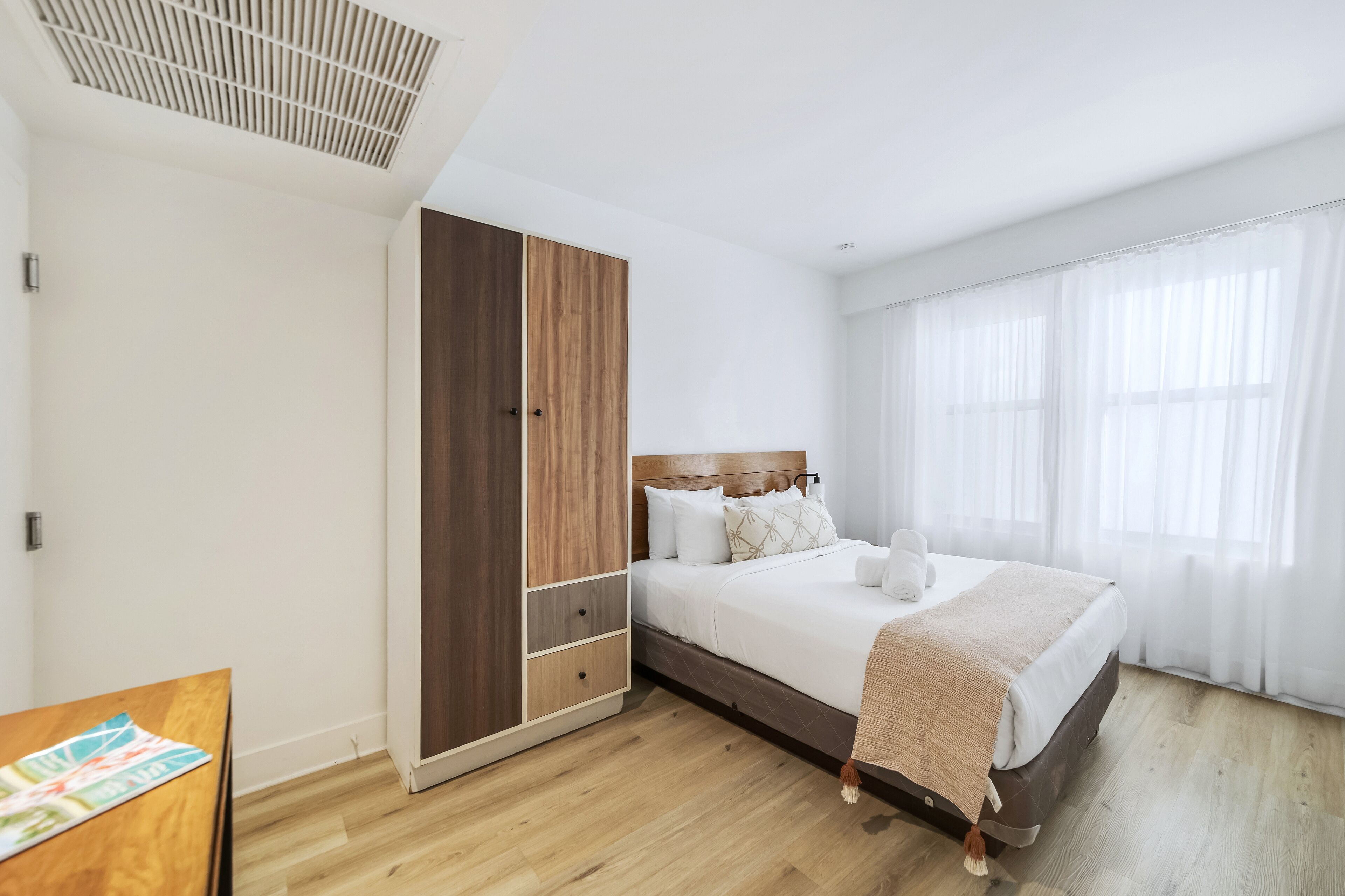A bedroom with a bed, wooden wardrobe, and a window with sheer curtains.