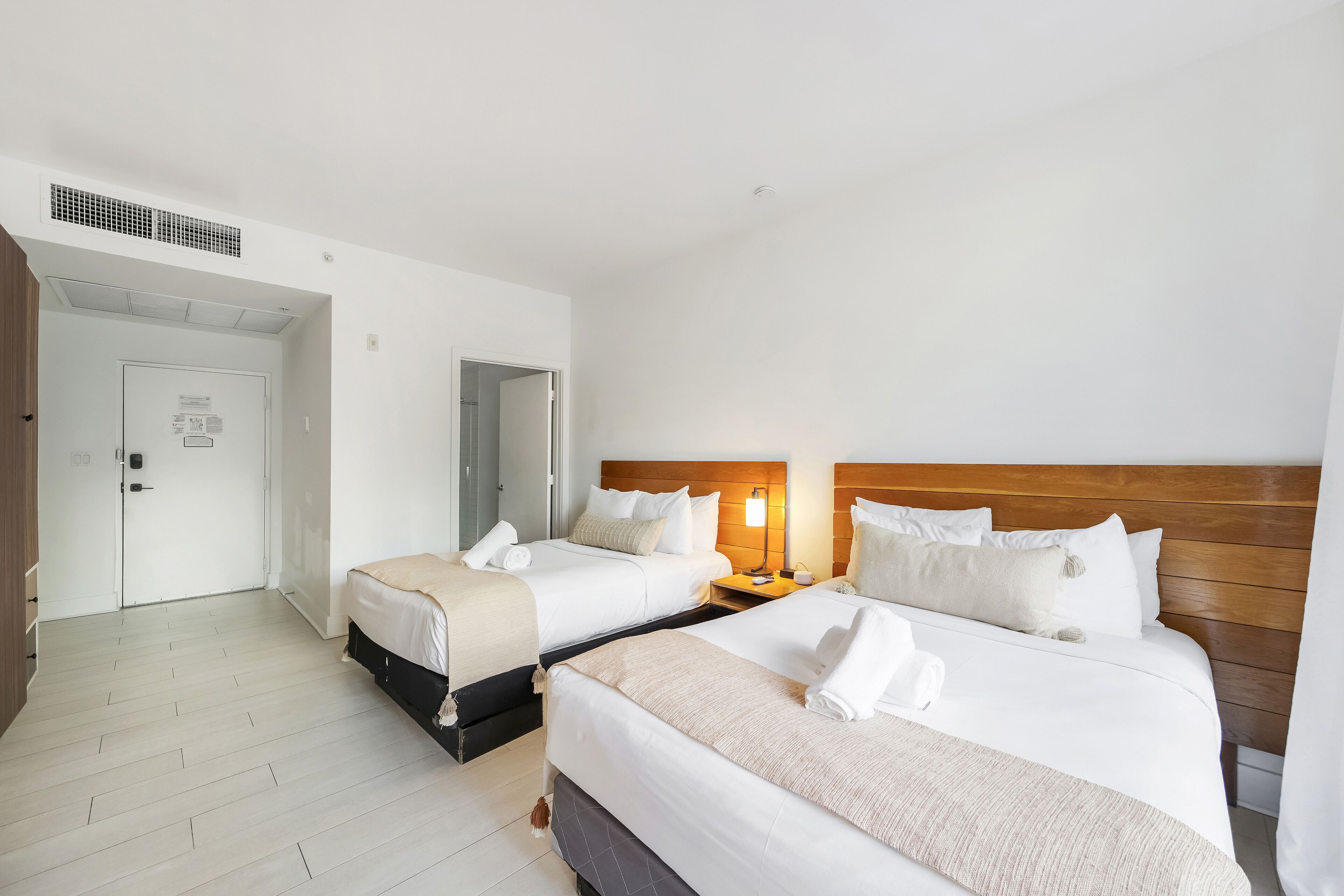 A hotel room with two beds, a wooden headboard, a bedside table with a lamp, and a door with a sign.