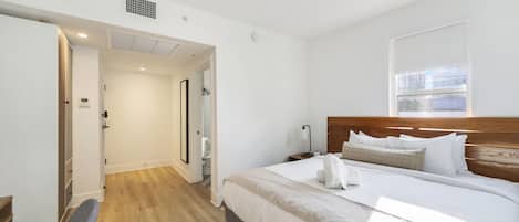 Une chambre d’hôtel moderne avec un plancher en bois, un grand lit avec une literie blanche, une tête de lit en bois et une fenêtre avec des stores.
