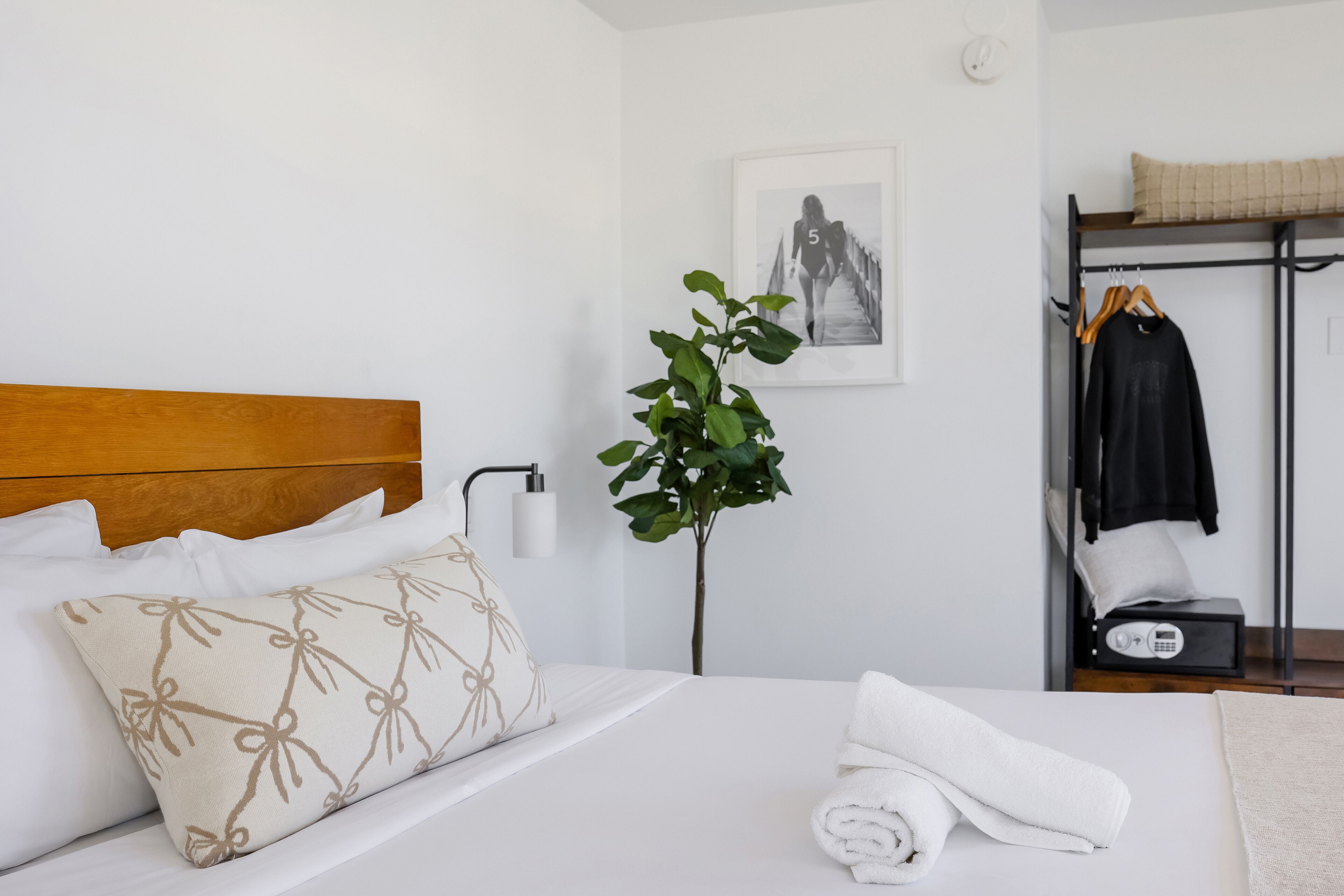 A neatly made bed with white linens and a decorative pillow, a bedside plant, a framed picture on the wall, and a clothing rack with a jacket and a pillow.