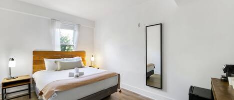 Habitación de hotel moderna con cama de cabecera de madera, una mesita de noche con lámpara, un espejo grande y un escritorio con cafetera.