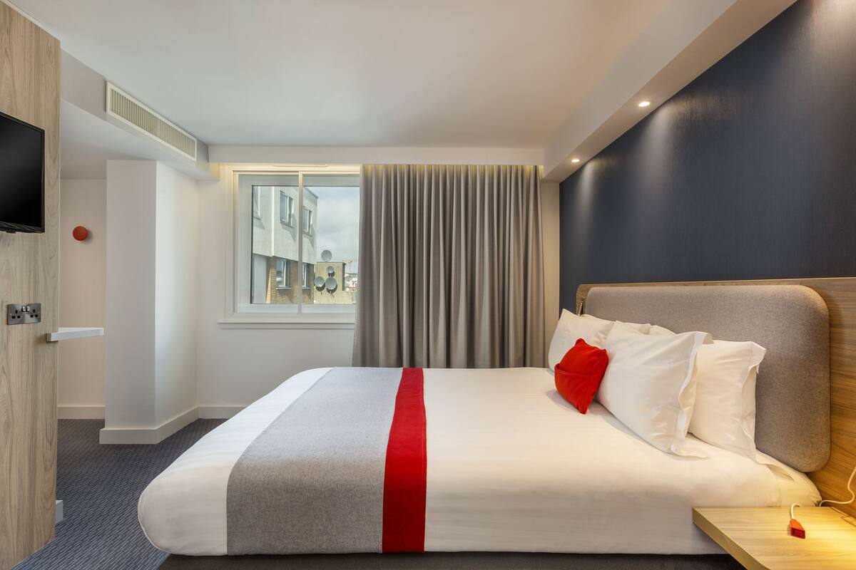 A hotel room with a large bed, a TV mounted on the wall, a window with curtains, and a view of buildings outside.