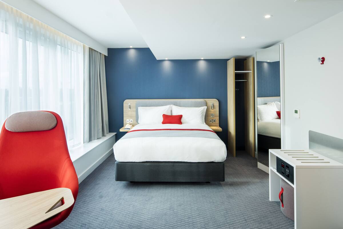 A modern hotel room with a large bed, a red chair, a desk, and a wardrobe.