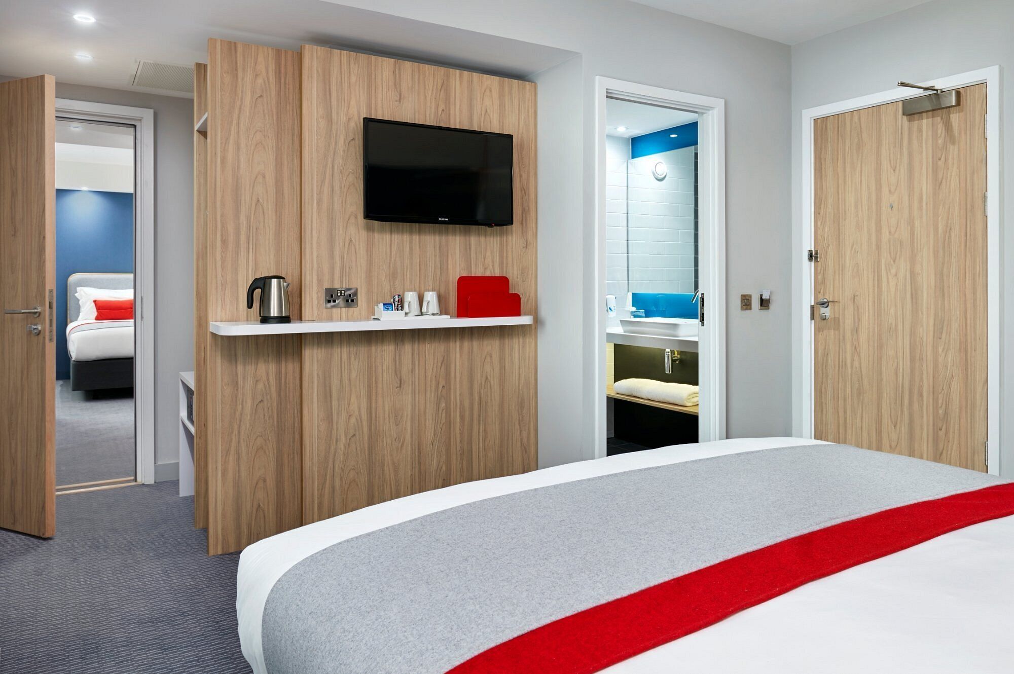 Habitación de hotel con cama, televisor montado, cafetera y estante con artículos de tocador.