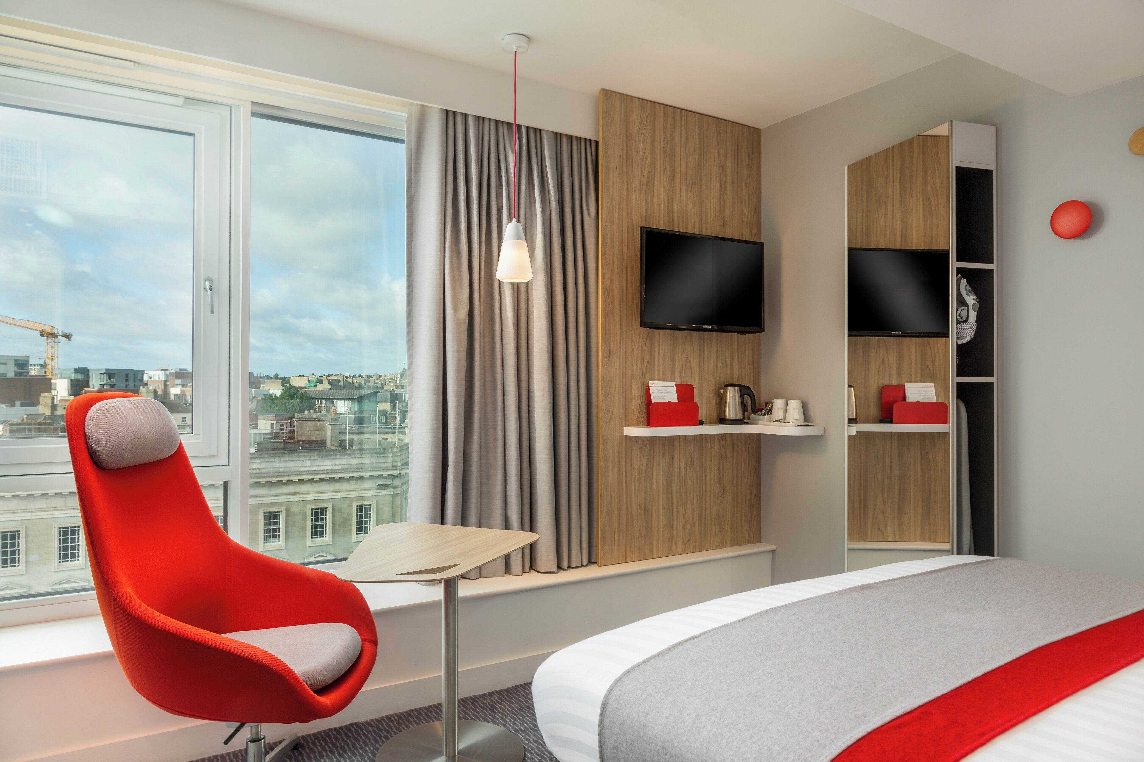 A hotel room with a large window, a red chair, a bed, a TV, and a shelf with items.