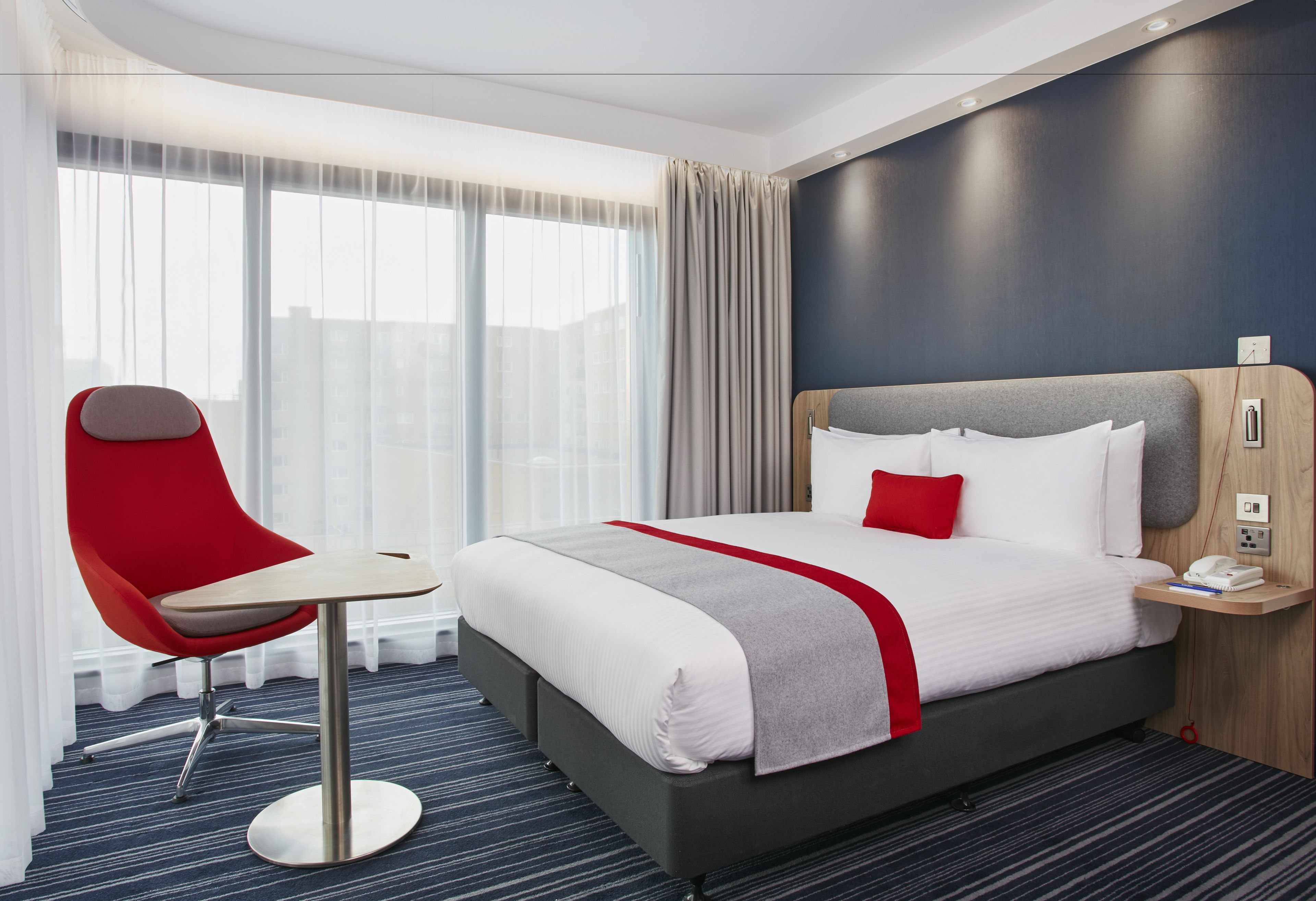 A hotel room with a large bed, a red chair, a bedside table, and a view of the city through large windows.