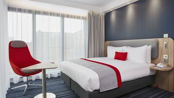 A hotel room with a large bed, a red chair, a bedside table, and a view of the city through large windows.