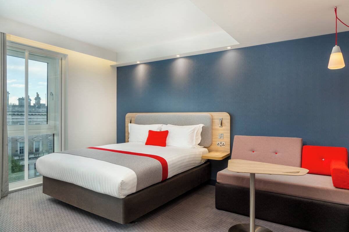 A modern hotel room with a large bed, a sofa, a small table, and a city view through the window.