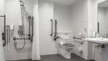 A modern bathroom with white tiles, a shower with a handheld showerhead, a toilet with a raised seat, and a sink with a mirror.