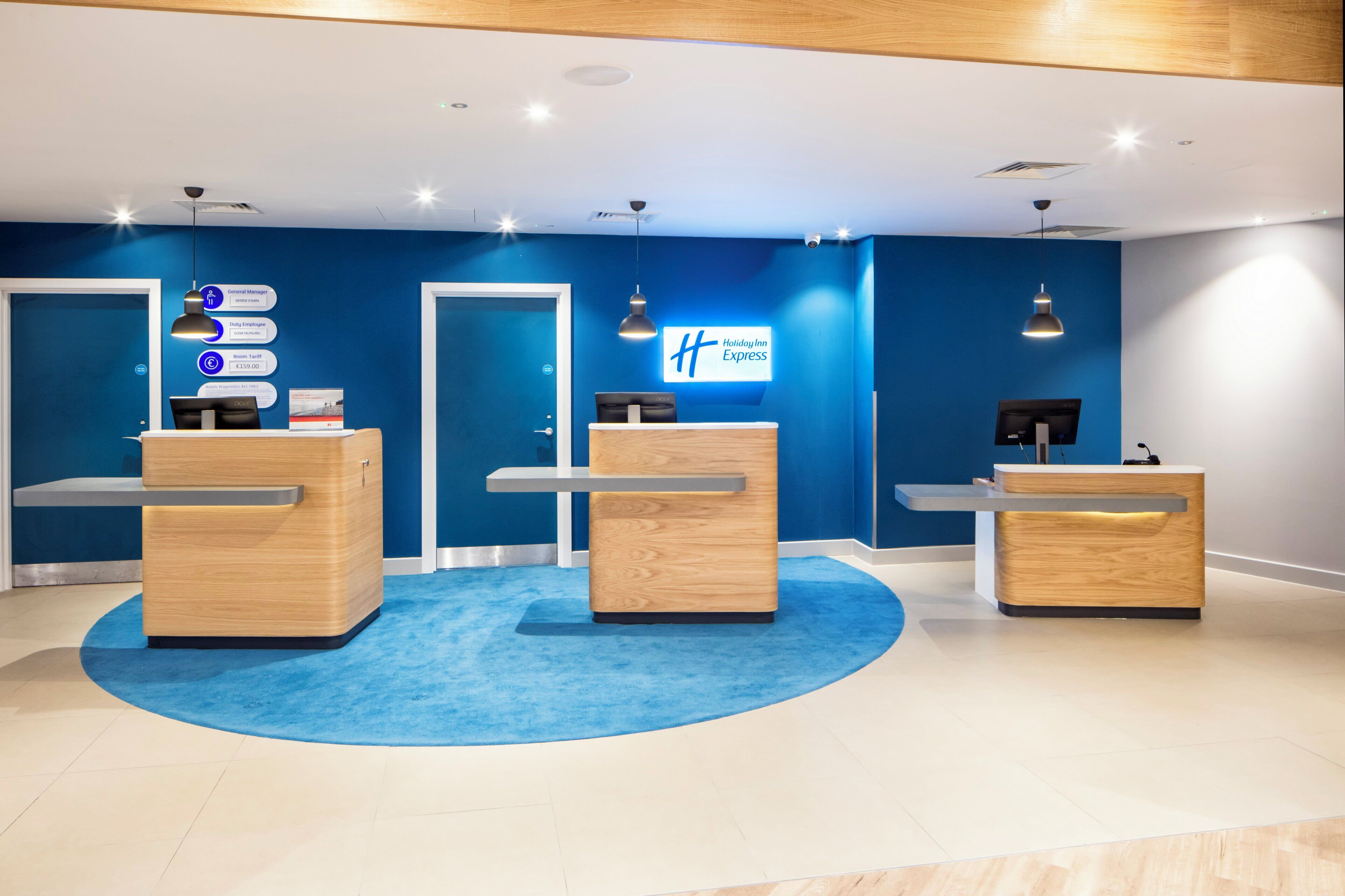 Aula, jossa on kolme puista vastaanottotiskiä, sininen matto ja seinällä Holiday Inn Express -kyltti.