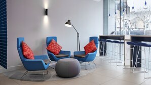 Ein moderner Wartebereich mit blauen Stühlen, roten Kissen, einem grauen Hocker und einer Stehlampe.